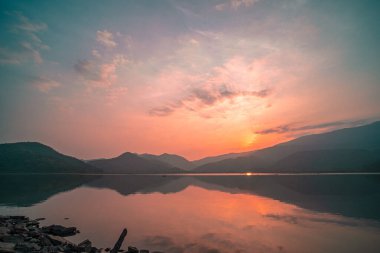 Güneş doğarken yansıması mükemmel olan dağ gölü manzarası. Pembe pastel gökyüzü, arka planda tepeler ve suya yansıyan güzel dağ manzarası. Doğa gölü manzarası