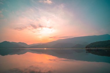 Güneş doğarken yansıması mükemmel olan dağ gölü manzarası. Pembe pastel gökyüzü, arka planda tepeler ve suya yansıyan güzel dağ manzarası. Doğa gölü manzarası