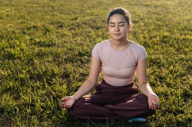 Asyalı görünümlü genç bir kız çimenlerde oturur ve meditasyon yapar..