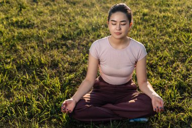 Asyalı görünümlü genç bir kız çimenlerde oturur ve meditasyon yapar..