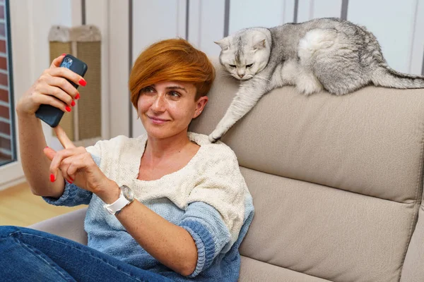 Çilli bir kız kanepeye oturur ve kanepenin arkasında yatan İngiliz bir kediyle selfie çeker..