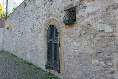 An ancient stone wall with a door and a small window, closed with wrought iron bars. Part of a cobbled pedestrian area.