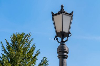 Mavi gökyüzünün arka planına ve yeşil bir ağacın tacına karşı sokak feneri. Kapat..