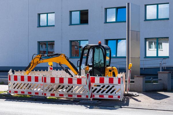 Yellow excavator fenced with traffic barriers. Light building with windows in the background.