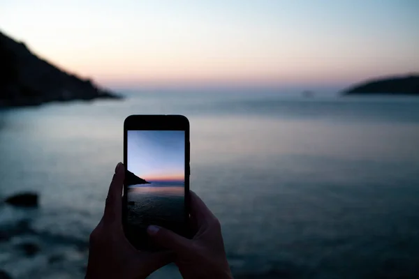 女子手牵着手机在日出时分在海湾上空 用手机拍摄落日在岩石上的照片 有选择地关注智能手机 免版税图库图片