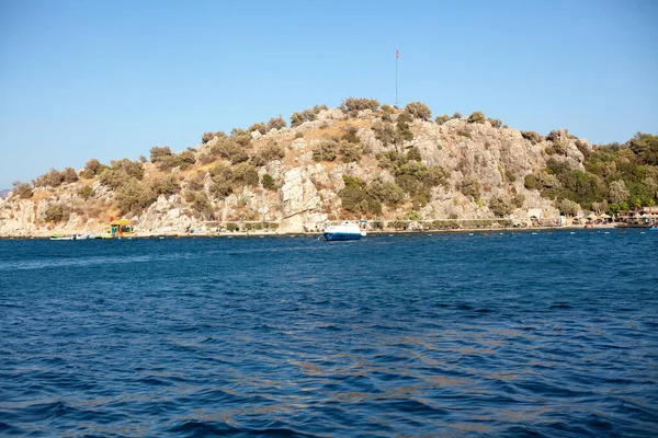 Kamień w zatoce morskiej z piaszczystą plażą i turecką flagą na szczycie w małej turystycznej miejscowości Turunc. Krajobraz z cumowanym jachtem rekreacyjnym. — Zdjęcie stockowe