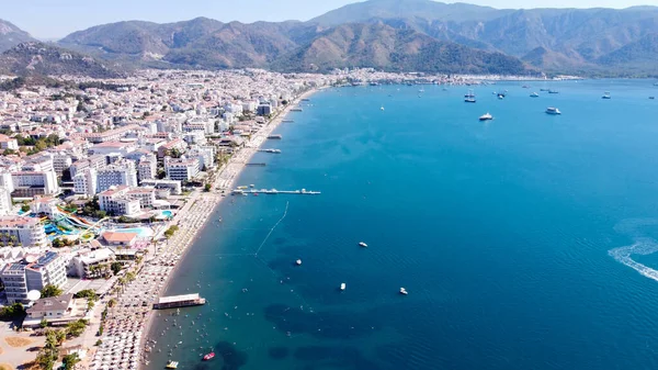 Türkiye 'de kalabalık plajlı, güneşli bir Marmaris şehrinin havadan görünüşü. Mavi deniz yüzeyinde tekne izleri var. Beyaz kumlu kumsal manzarası