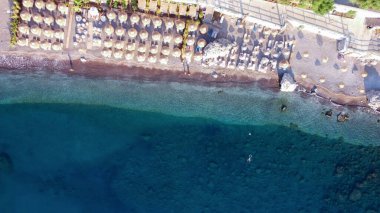 Sabahları boş bir sahilin havadan görünüşü, saman şemsiyeleri, denizde yüzen insanlar. Çakıl taşı plajının drone görüntüsü. Altında taşlar olan şeffaf deniz suyu..