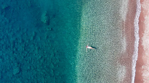 Egy fiatal, csinos nő egy fehér, egyrészes fürdőruhában, amint türkiz tengervízben borotválkozik. A nő a vízátlátszó vízen fekszik. Aeriale drónfotó. — Stock Fotó