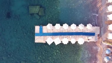 Plaj şemsiyeli ahşap iskele manzarası ve turistik tatil beldesinde güneşlik. Şeffaf deniz suyu. Deniz dibindeki kayalar. İnsansız hava aracı görüntüsüyle sakin deniz
