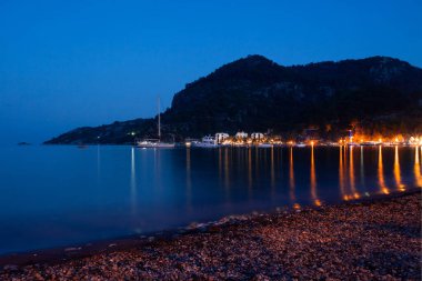 Night view of the embankment and promenade in the light of street lights and lighting of restaurants. small town surrounded by mountains in evening. Light reflections on sea water surface. Night beach