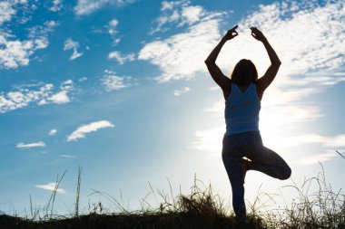 Sahildeki bir kumulda gün doğumunda güneş ışığıyla vrksasana yoga pozu veren bir kadının silueti.