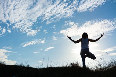 Sahildeki bir kumulda gün doğumunda güneş ışığıyla vrksasana yoga pozu veren bir kadının silueti.