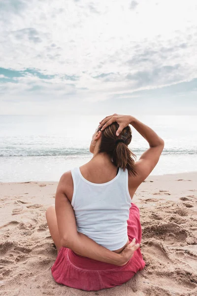 Sahilde Virasana Asana ya da Kahraman pozu veren bir kadın, arka planda deniz varken boyun esnetiyor.