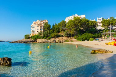 Calvia, Mallorca, İspanya. 18 Temmuz 2022 - Illetes plajında insanlar çam ağaçlarıyla çevrili, önünde Sa Caleta adası, sabahın erken saatlerinde