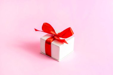 Açık pembe arka planda parlak kırmızı kurdeleli küçük beyaz paketlenmiş hediye kutusu. Sevgililer günü, doğum günü, anneler günü sürprizi hediyesi.