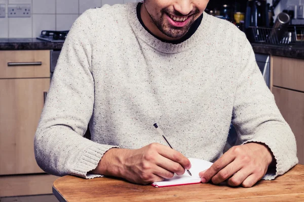 Uśmiechający się młody człowiek pisze w swojej kuchni — Zdjęcie stockowe