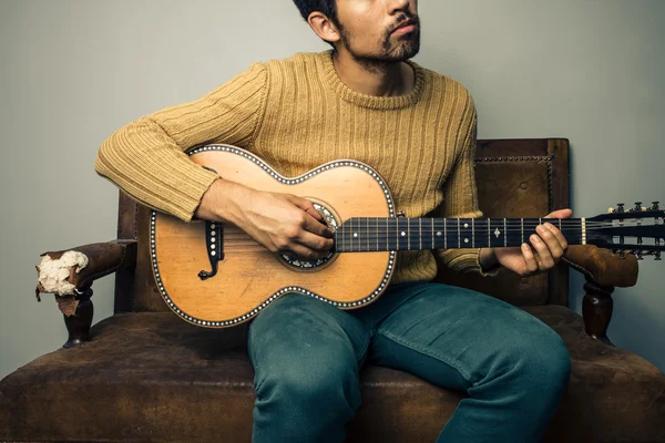 Młody człowiek na starej kanapie jest gra na gitarze — Zdjęcie stockowe