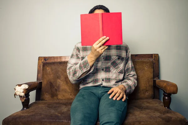 Homme caché derrière le livre sur un vieux canapé — Photo