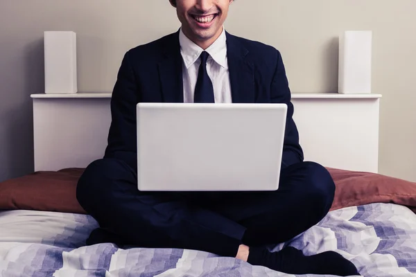 Feliz joven empresario sentado en la cama con el ordenador portátil —  Fotos de Stock