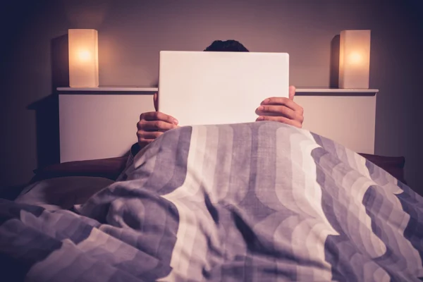 El hombre está sentado en la cama detrás de su portátil —  Fotos de Stock