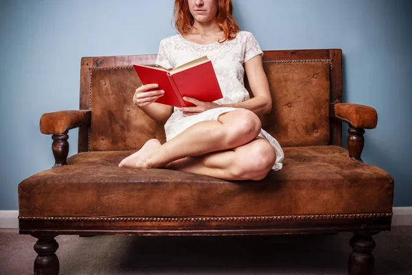 夏天穿裙子的年轻女子是坐在旧沙发上，阅读 — 图库照片