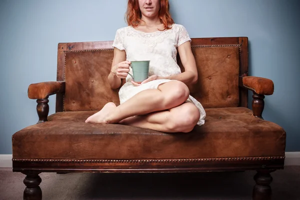 年轻女子坐在旧沙发上喝茶的夏天穿裙子 — 图库照片