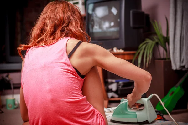 Jovem engomando suas roupas em casa e assistindo TV — Fotografia de Stock