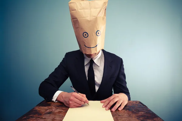 Businessman with bag over head writing at desk — Stock Photo, Image