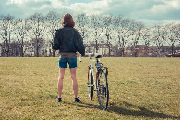 Ung kvinna som står i parken med cykel — Stockfoto