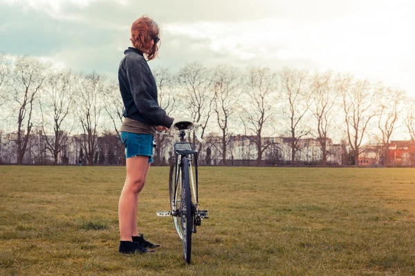 Ung kvinna som står i parken med cykel — Stockfoto