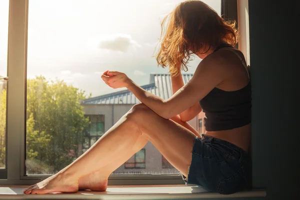 Ung kvinna sitter på fönsterbrädan solig dag — Stockfoto
