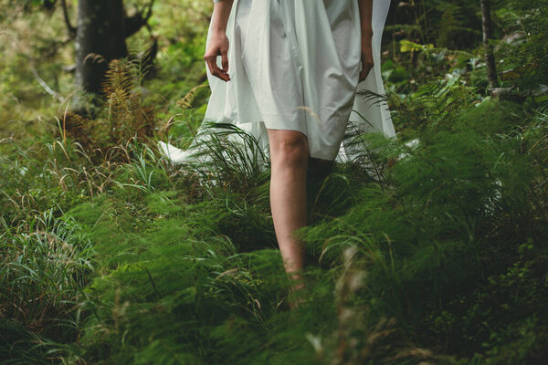 Legs of a young girl who walks barefoot in a white dress on green grass in the forest on a sunny day, front view close-up