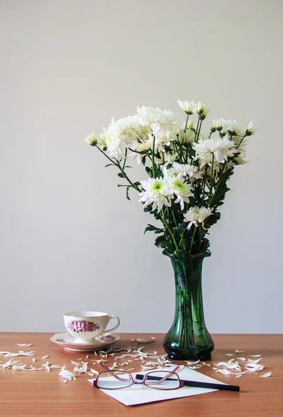 Romantisch ein Strauß Chrysanthemen weiß in grüner Glaskanne mit Kaffeetasse, Bleistift, Gläsern und Umschlag auf Holztisch und grauen Betonwänden — Stockfoto