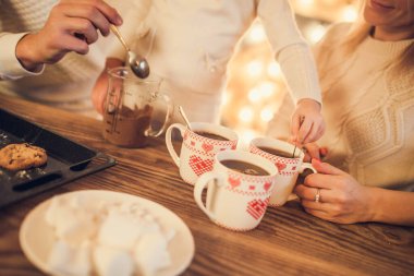 Anne, baba ve beyaz kazaklı kız şekerli kakao pişirir ve içerler. Ellerinizi ve bardaklarınızı yakın tutun. Noel konsepti.