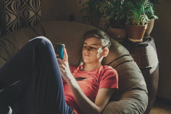 Een Jongeman Kijkt Naar Zijn Smartphone Liggend Een Stoel Thuis — Stockfoto