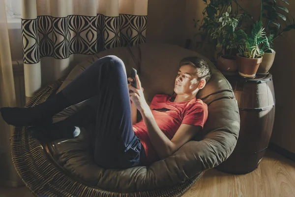 Een Jongeman Kijkt Naar Zijn Smartphone Liggend Een Stoel Thuis — Stockfoto