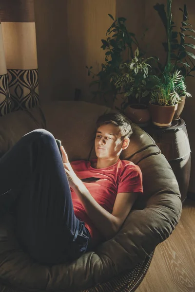 Een Jongeman Kijkt Naar Zijn Smartphone Liggend Een Stoel Thuis — Stockfoto