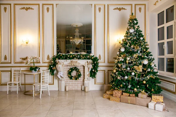 Classic apartments hall with a white fireplace, decorated christmas tree with gift boxes, large mirror and windows.