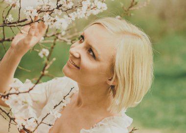 Beautiful young woman in a white chiffon dress with a short blonde haircut looks at a blooming cherry branch, admires and rejoices in spring.