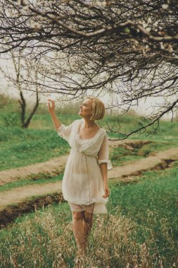 Beautiful young woman in a white chiffon dress with a short blonde haircut looks at a blooming cherry branch, admires and rejoices in spring