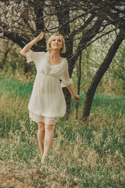 Beautiful young woman in a white chiffon dress with a short blonde haircut looks at a blooming cherry branch, admires and rejoices in spring