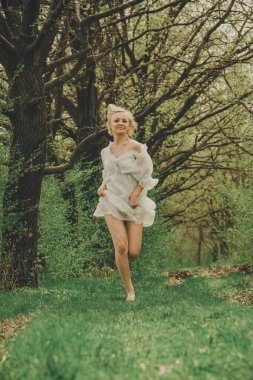 Beyaz şifon elbiseli, kısa sarı saçlı güzel bir kadın parkta koşuyor..