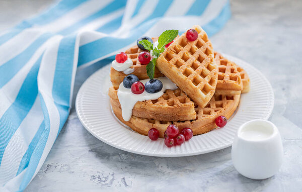 Waffles with blueberries, red currants, poured with yogurt on a white porcelain plate.