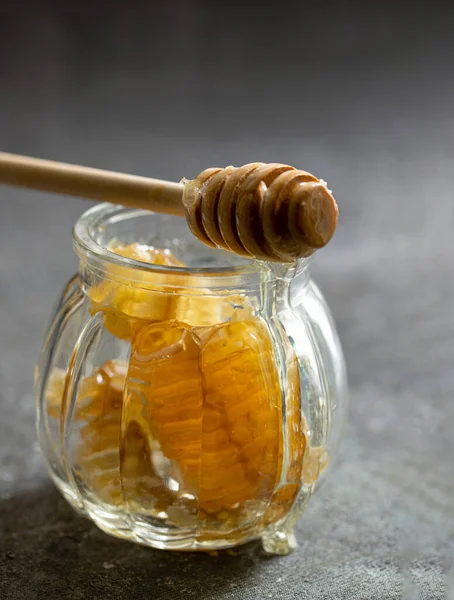 Peigne de miel scellée avec trempette à miel en bois pour mélanger et déguster du miel naturel dans un petit bocal en verre sur une table grise. — Photo