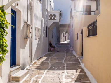 Ios, Greece - July 31, 2021: A typical greek village street scene with white wall and a old woman sitting in the shade. Taken at the end of a sunny summer day.