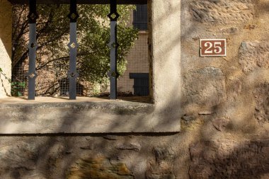 Street number twenty-five in the Southern France village of Alet-les-bains with white characters on a brown background, placed on a stone wall with foliage shadows