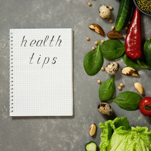 Diet plan with greens and fresh vegetables in a paper notebook on a gray table surface.