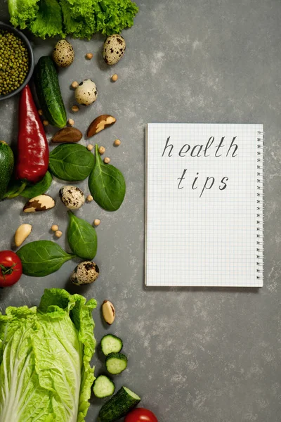 Diet plan with greens and fresh vegetables in a paper notebook on a gray table surface.