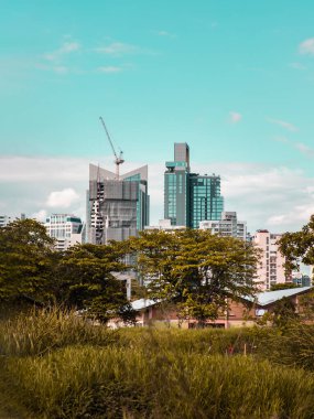 Bangkok, Tayland - 19 Şubat 2022: Doğada yeşil ağaçlar arasında yer alan modern yüksek binaların görüntüsü öğleden sonra bulutlu mavi gökyüzüne karşı. Şehir büyüme konsepti, Seçici odak.